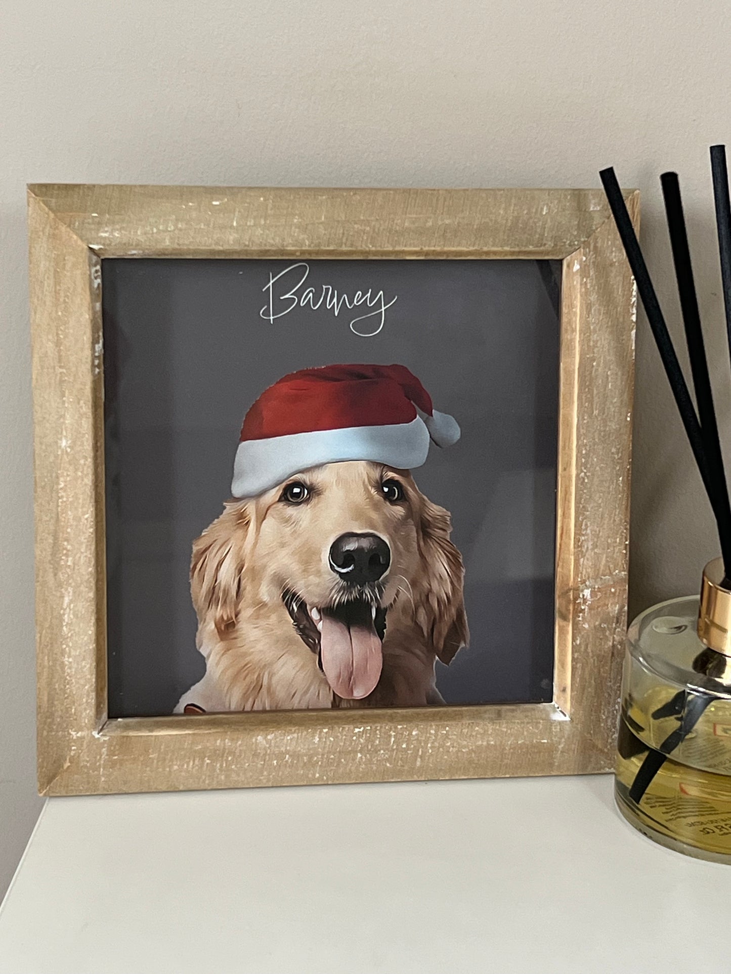 Framed picture of a dog wearing a Santa hat with 'Barney' written above, in a neutral box frame on a light-colored wall.