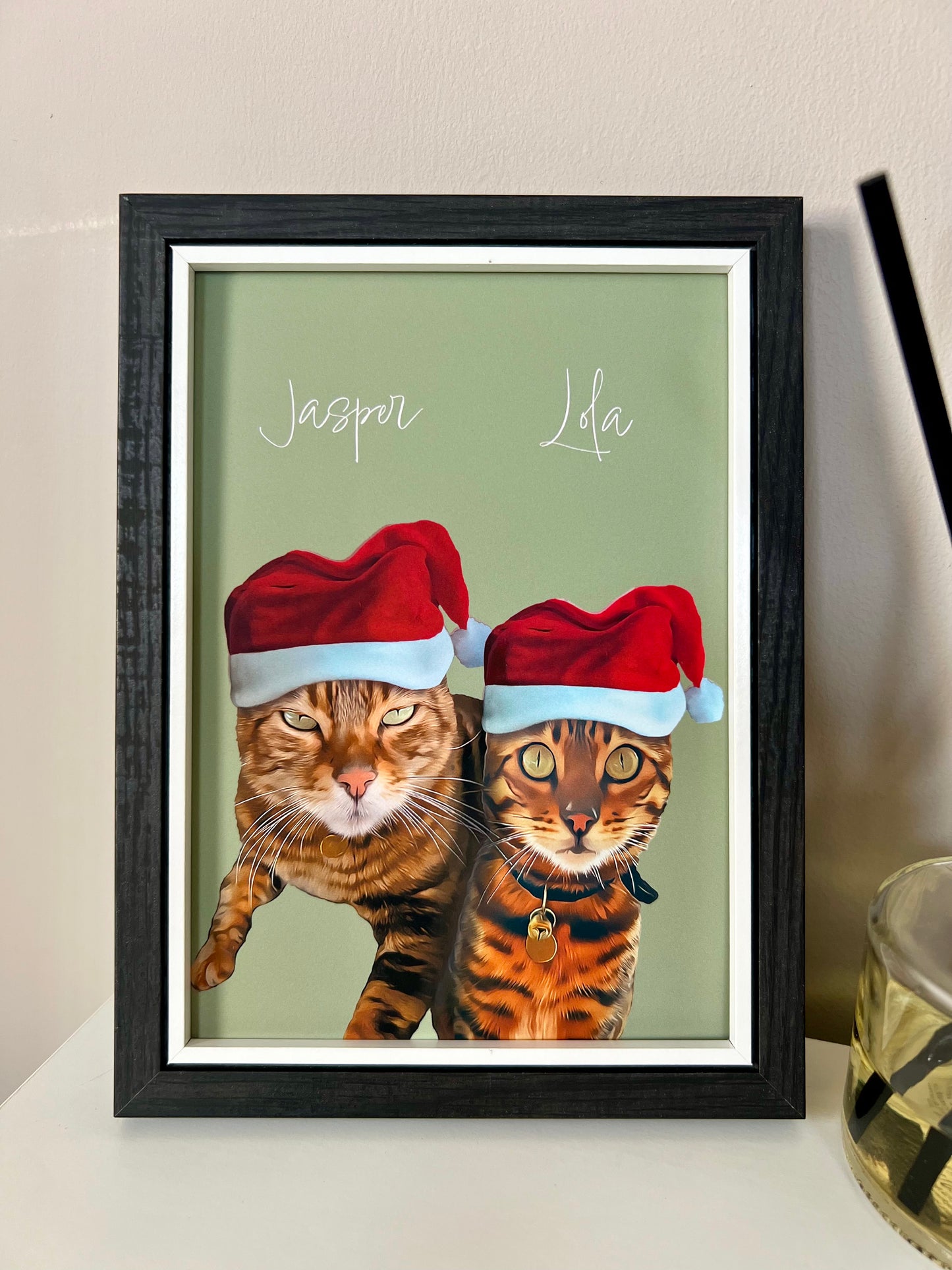 Framed artwork of two Bengal Cats wearing Santa hats in a black frame on a beige wall.