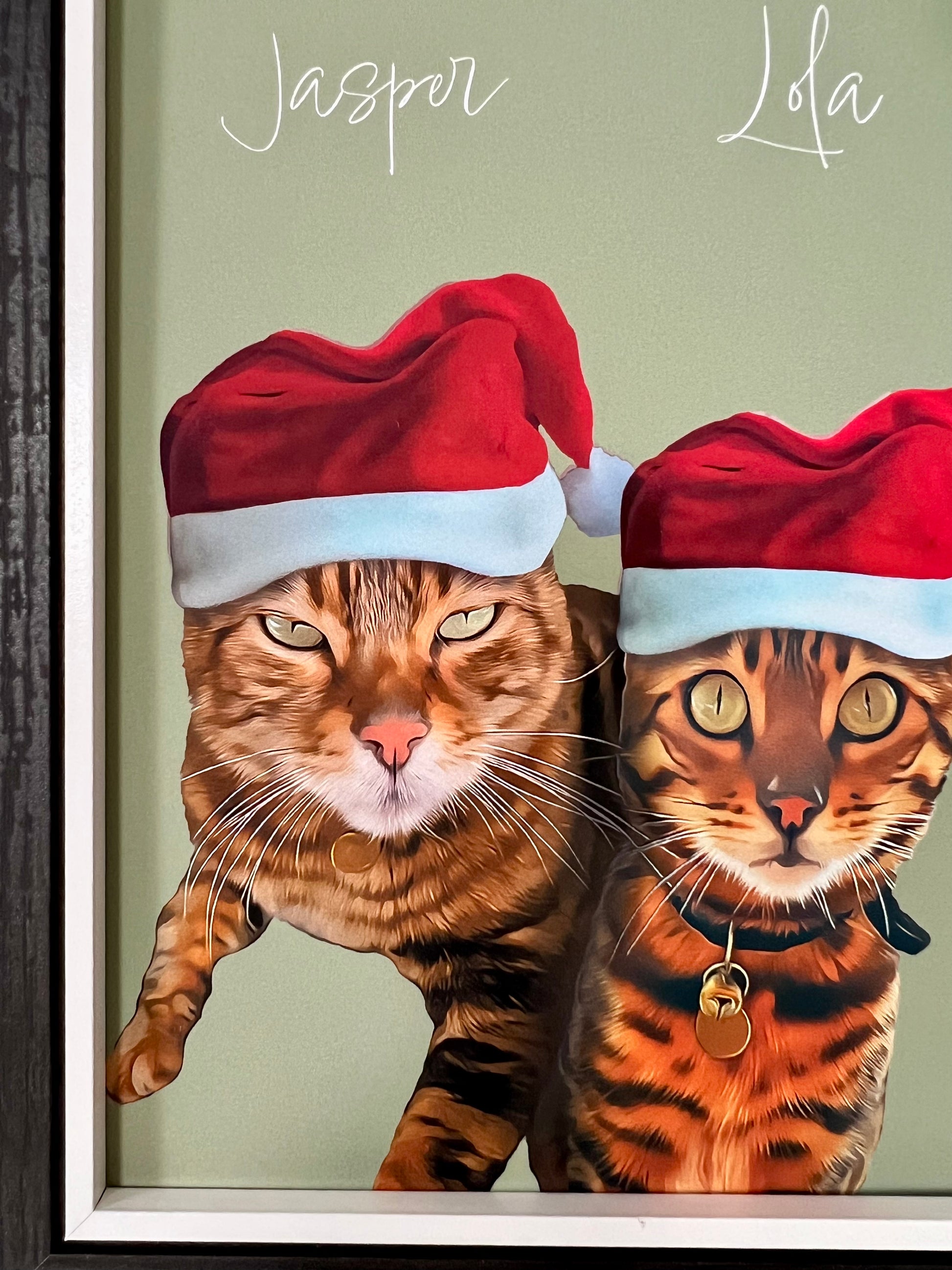 Close up left of framed artwork of two Bengal Cats wearing Santa hats in a black frame.