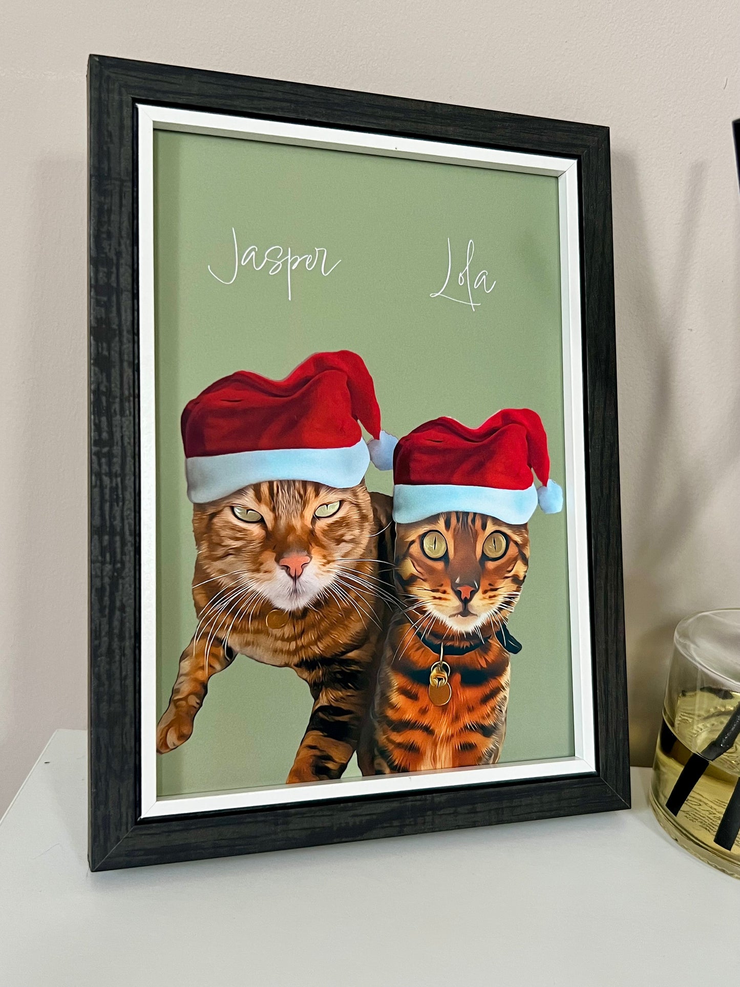 Side view of a Framed artwork of two Bengal Cats wearing Santa hats in a black frame on a beige wall.
