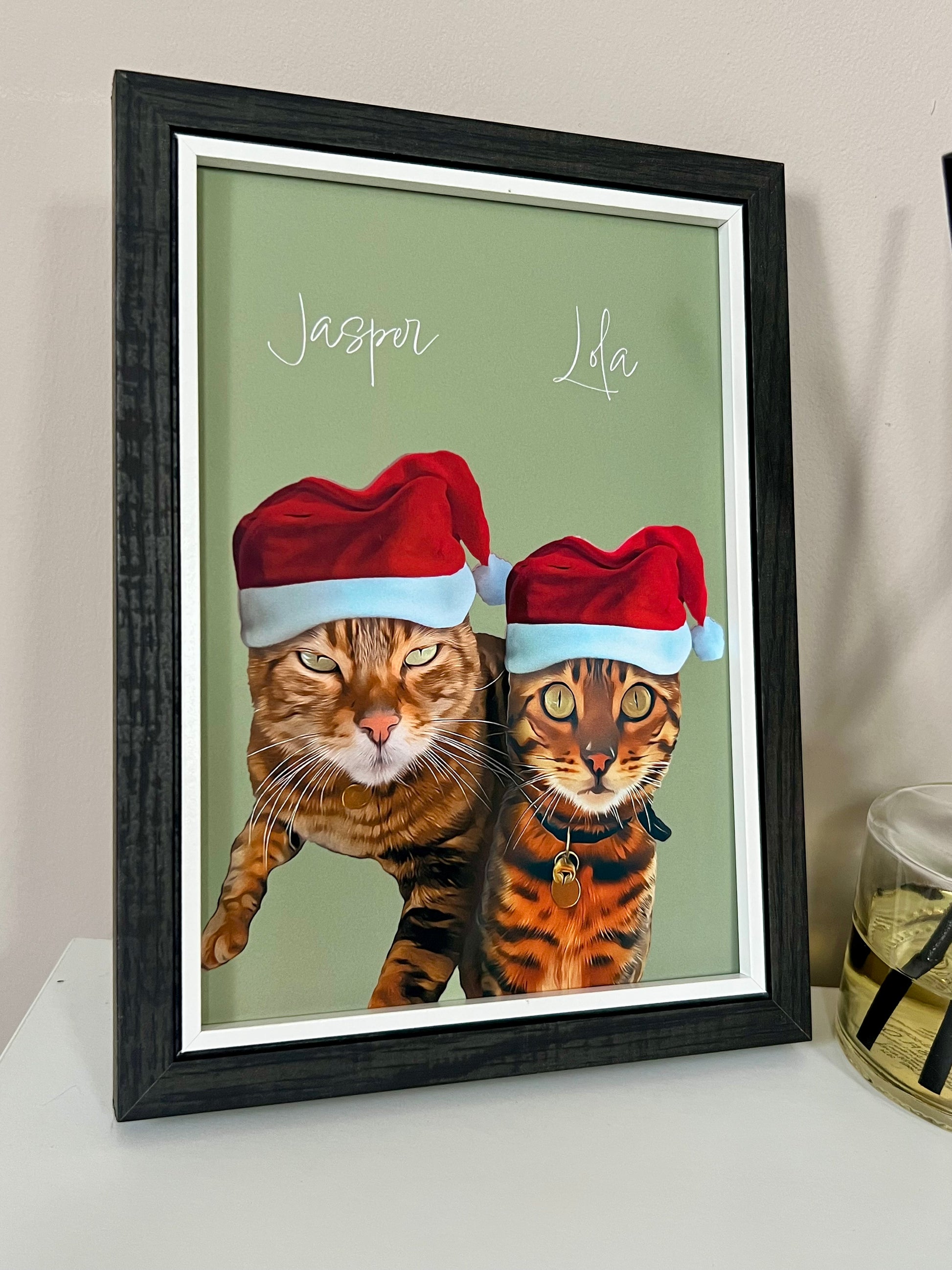 Side view of a Framed artwork of two Bengal Cats wearing Santa hats in a black frame on a beige wall.