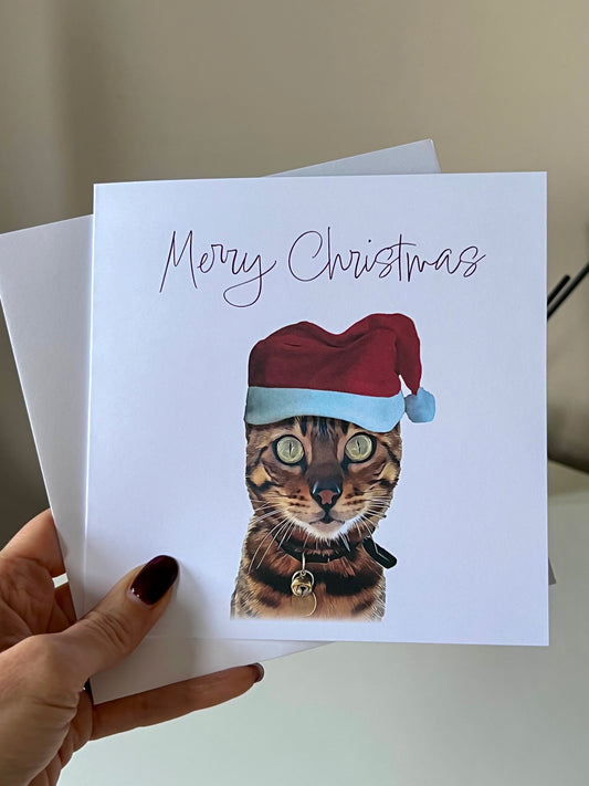 Custom Christmas Card of a Bengal Cat wearing a Santa hat with the words "Merry Christmas" held by and hand against a beige wall.