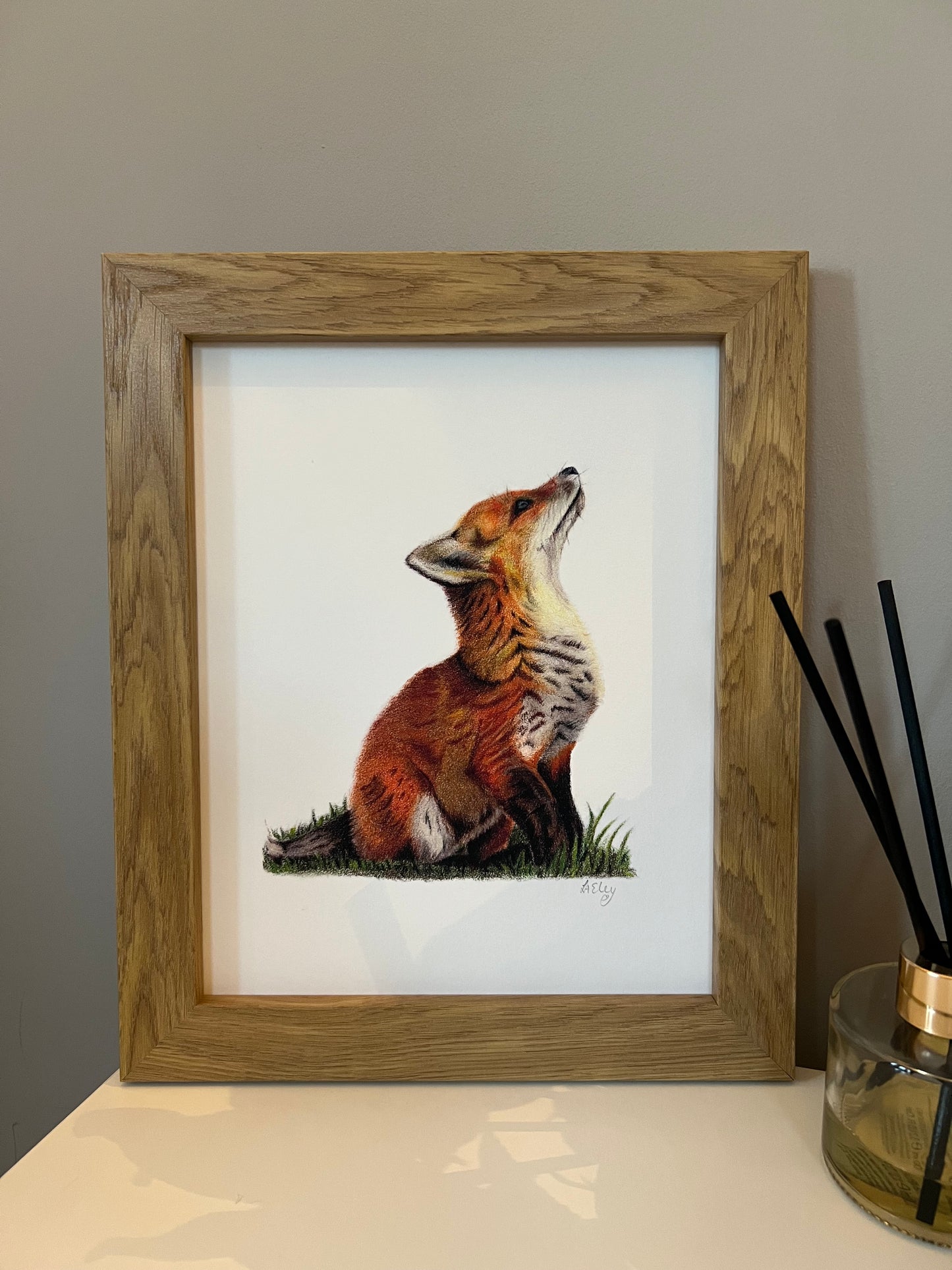 Framed artwork of a fox cub in vibrant oranges and yellows on a white background in a wooden oak style frame. 