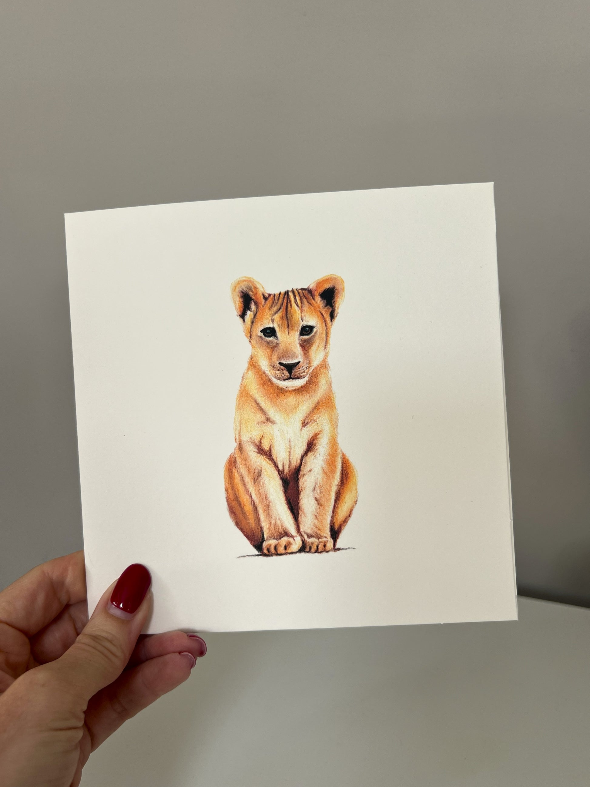 A hand with red nail polish holds a square white art greeting card featuring a detailed illustration of a sitting lion cub, set against a plain gray background.