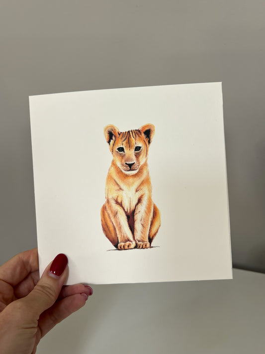 A hand with red nail polish holds a square white art greeting card featuring a detailed illustration of a sitting lion cub, set against a plain gray background.
