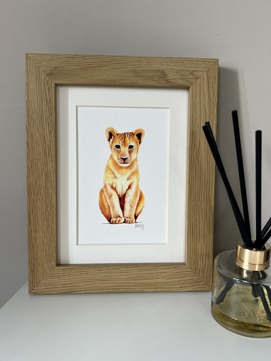 A framed picture of a lion cub sitting down, set in a light oak frame with a white mat. The framed print is styled on a white surface next to a reed diffuser against a neutral gray wall.