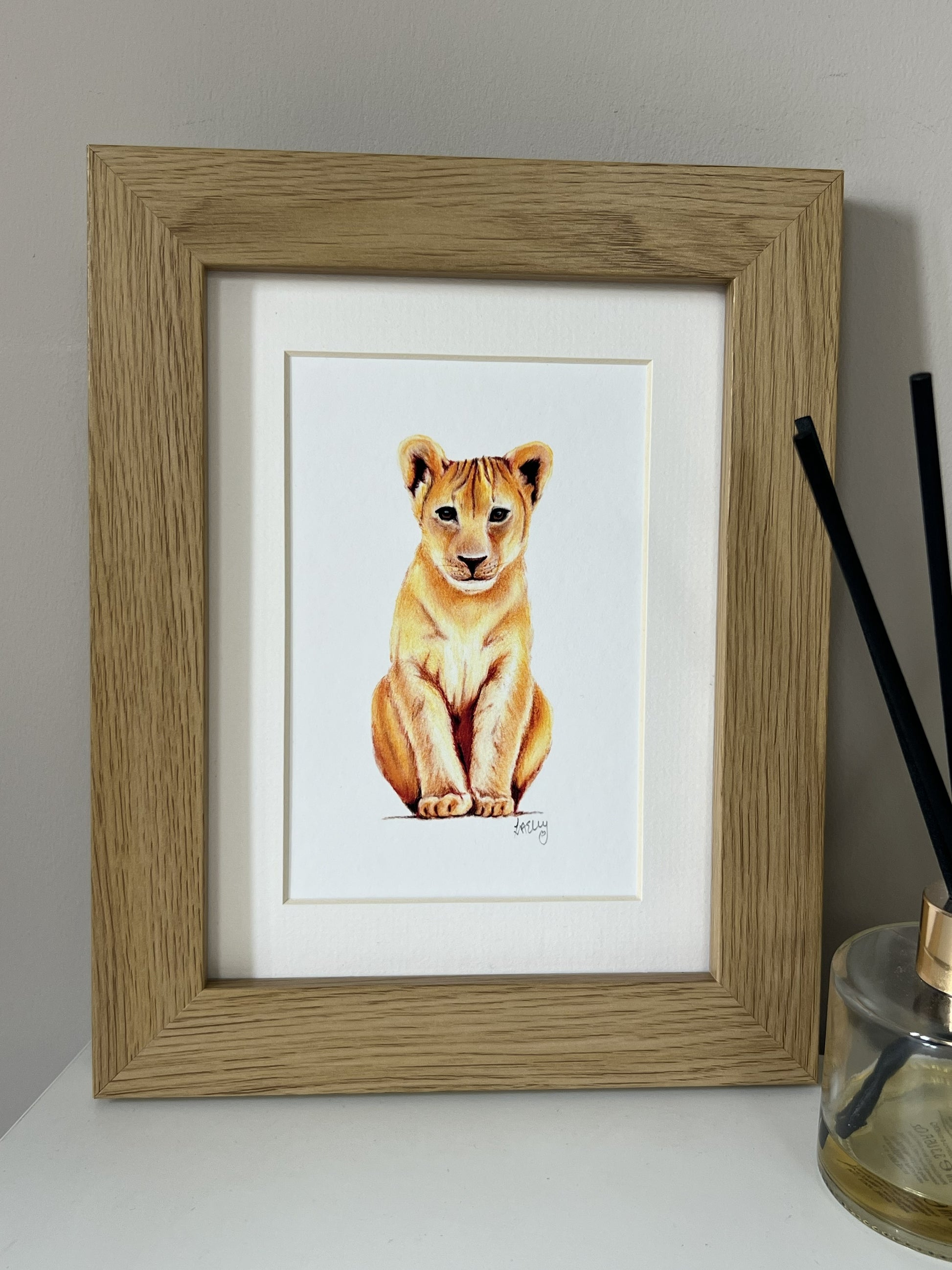 A framed lion cub drawing in a light oak frame with a white mount, displayed on a white surface next to a glass reed diffuser.