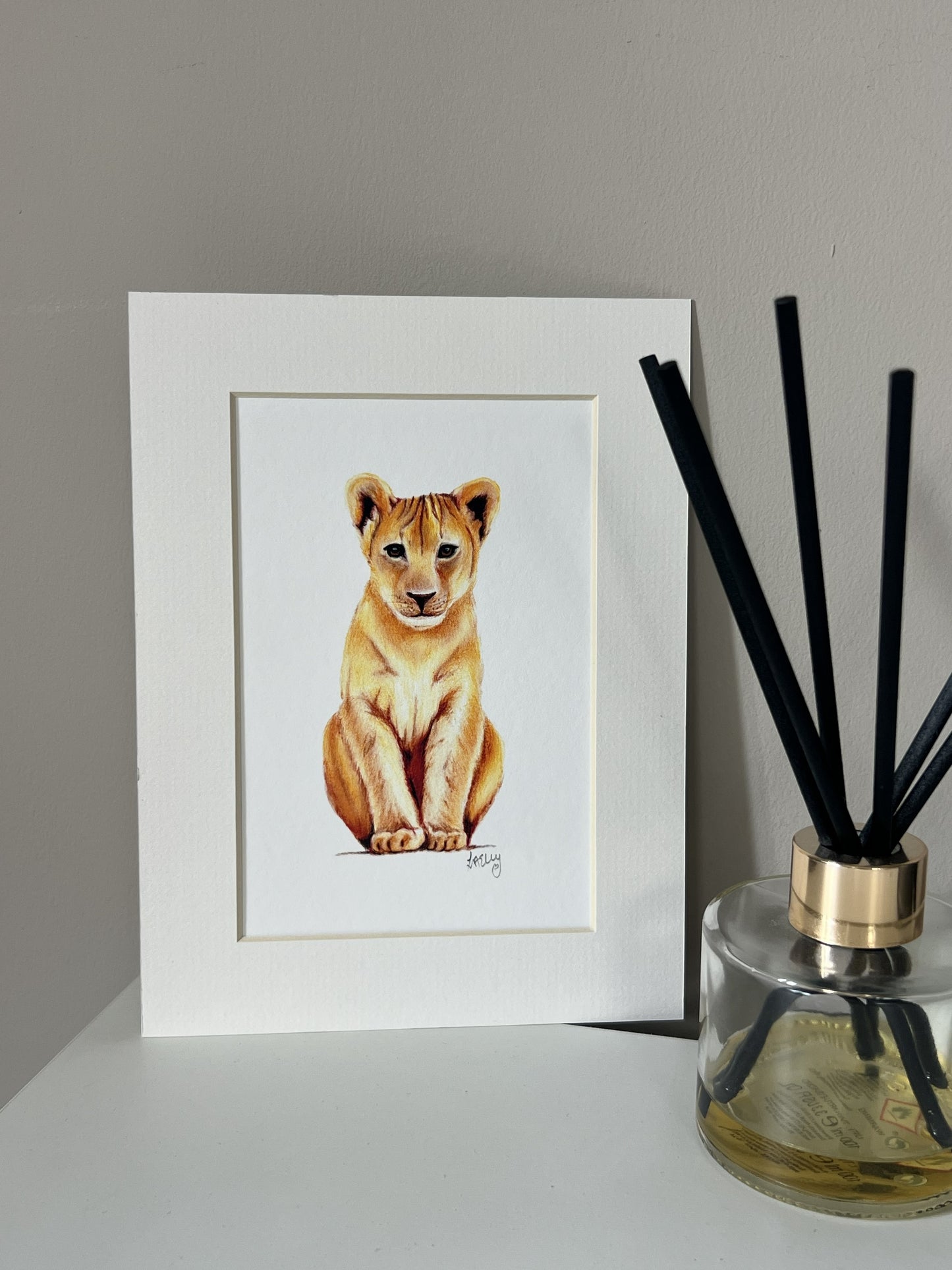A framed picture of a lion cub sitting down, displayed on a white surface next to a reed diffuser with black sticks.