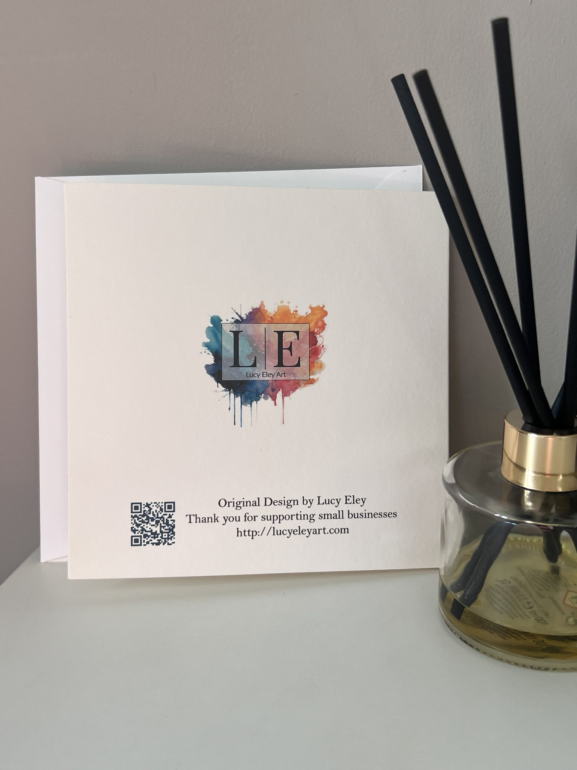 A white printed card back standing on a tabletop beside a glass reed diffuser bottle with black straws. The card has a colorful watercolor-style 'LE' Art logo and small printed text, photographed against a plain light wall.