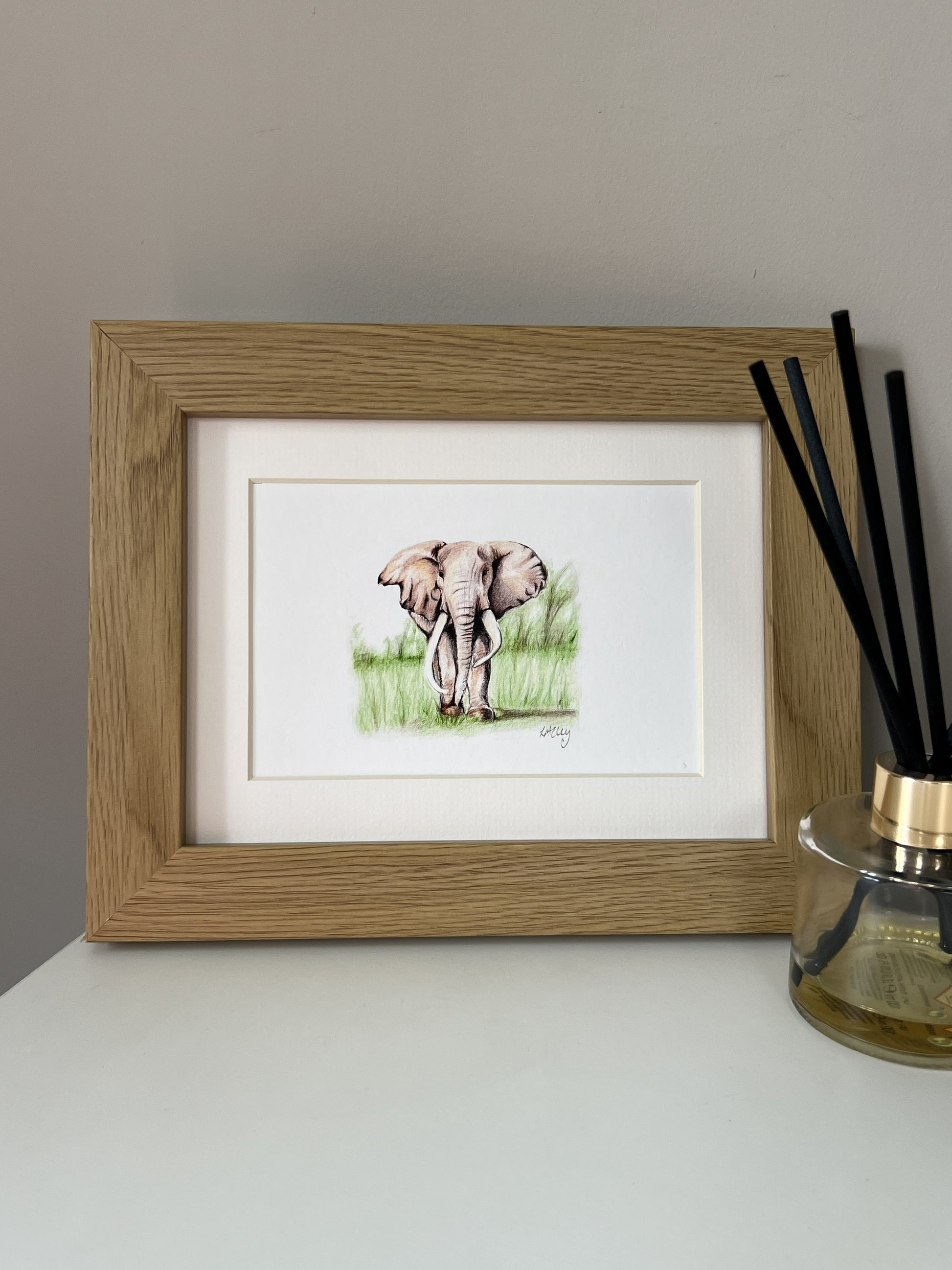 A framed pencil illustration of an elephant standing in grass, displayed on a white surface next to a glass reed diffuser against a neutral gray wall.