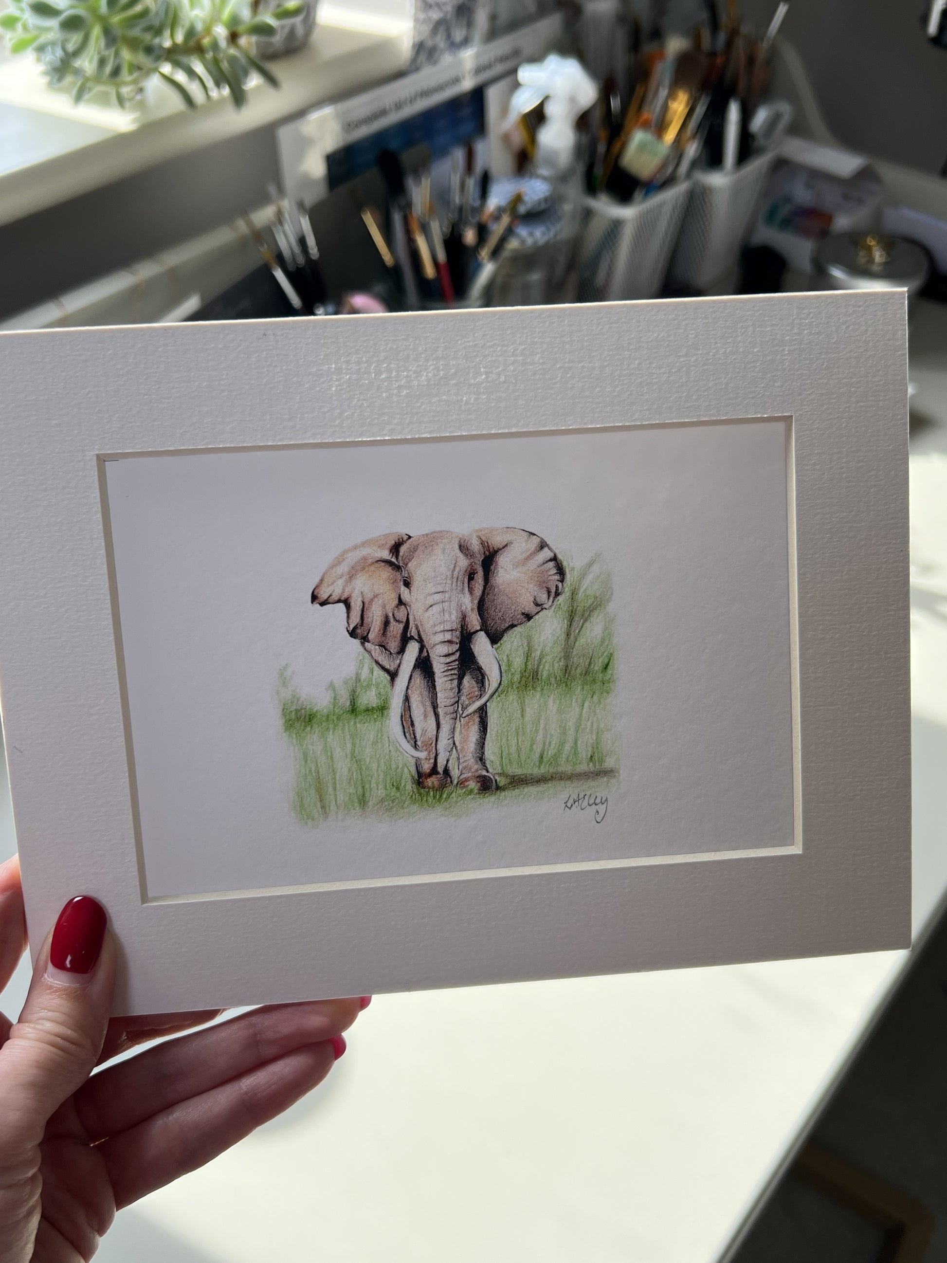 A hand with red nail polish holds a matted art print featuring a pencil illustration of an elephant standing in grass. The print has a wide white border and is photographed in a real indoor setting with a desk and art supplies blurred in the background.