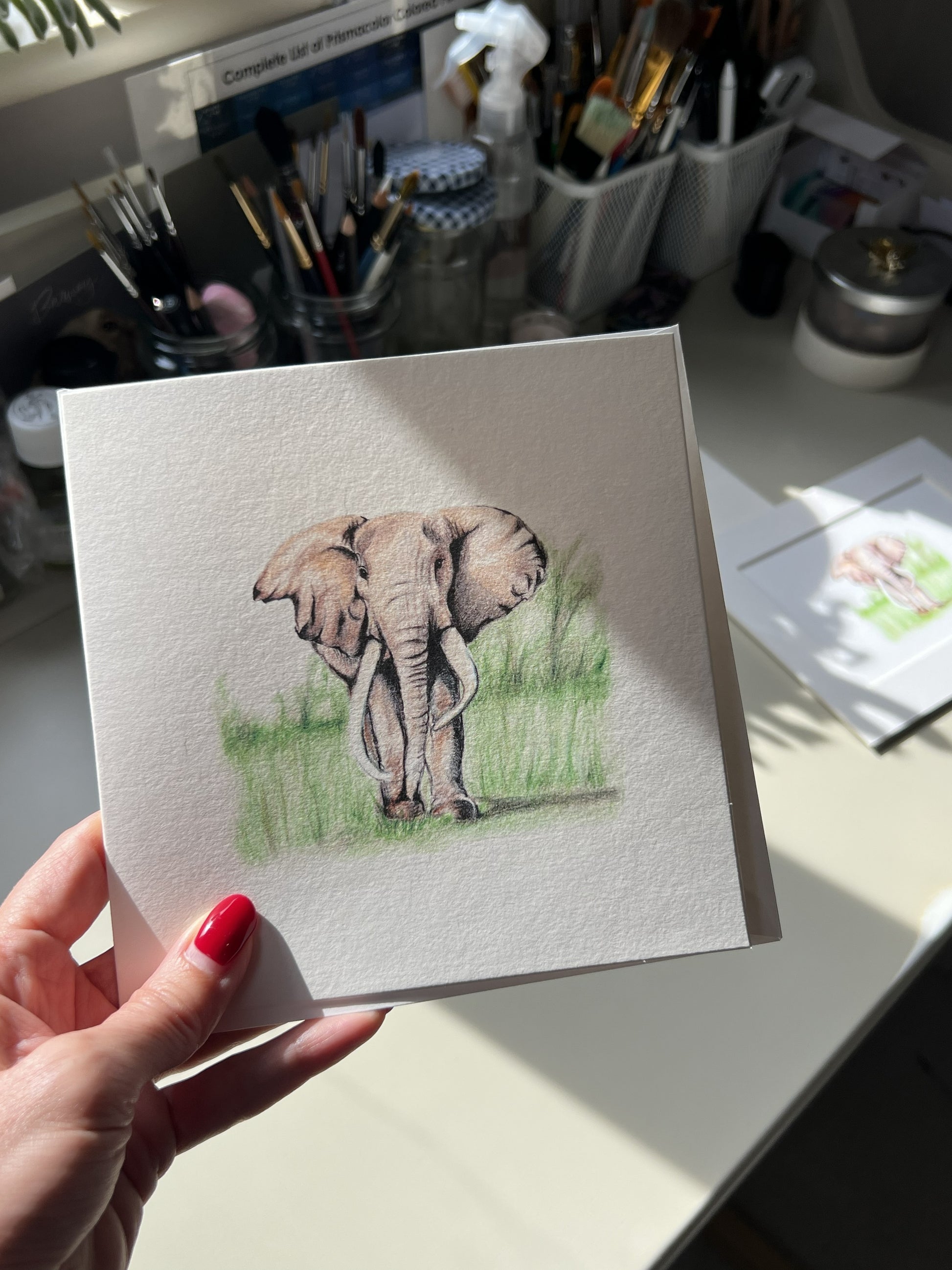 A hand with red nail polish holds a square greeting card pencil illustration of an elephant standing in green grass, photographed in a sunlit art studio or desk area with brushes and supplies in the background.