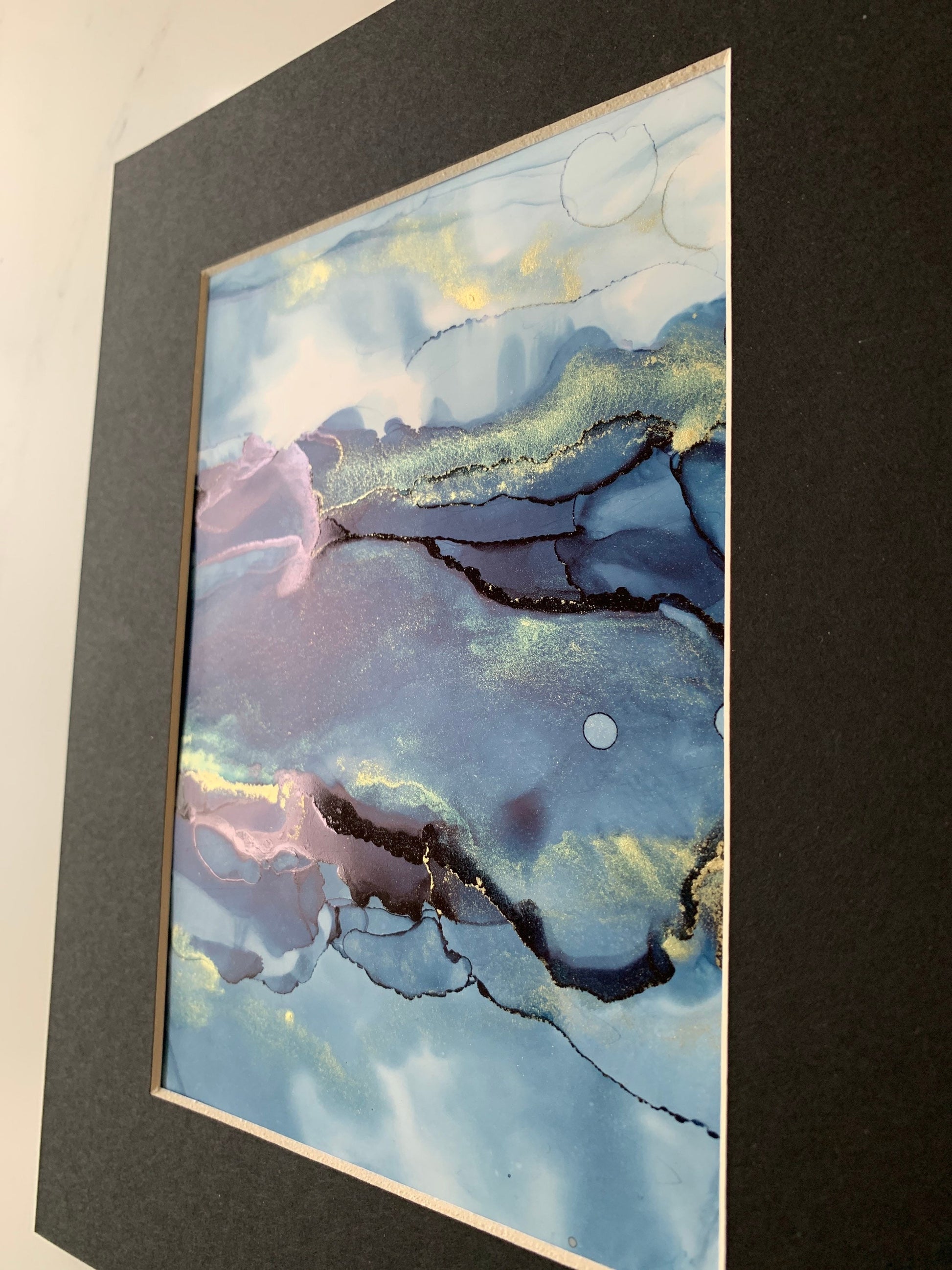 An A5 size contemporary ink painting featuring blue and metallic colours with gold shimmers, presented in a black mount.