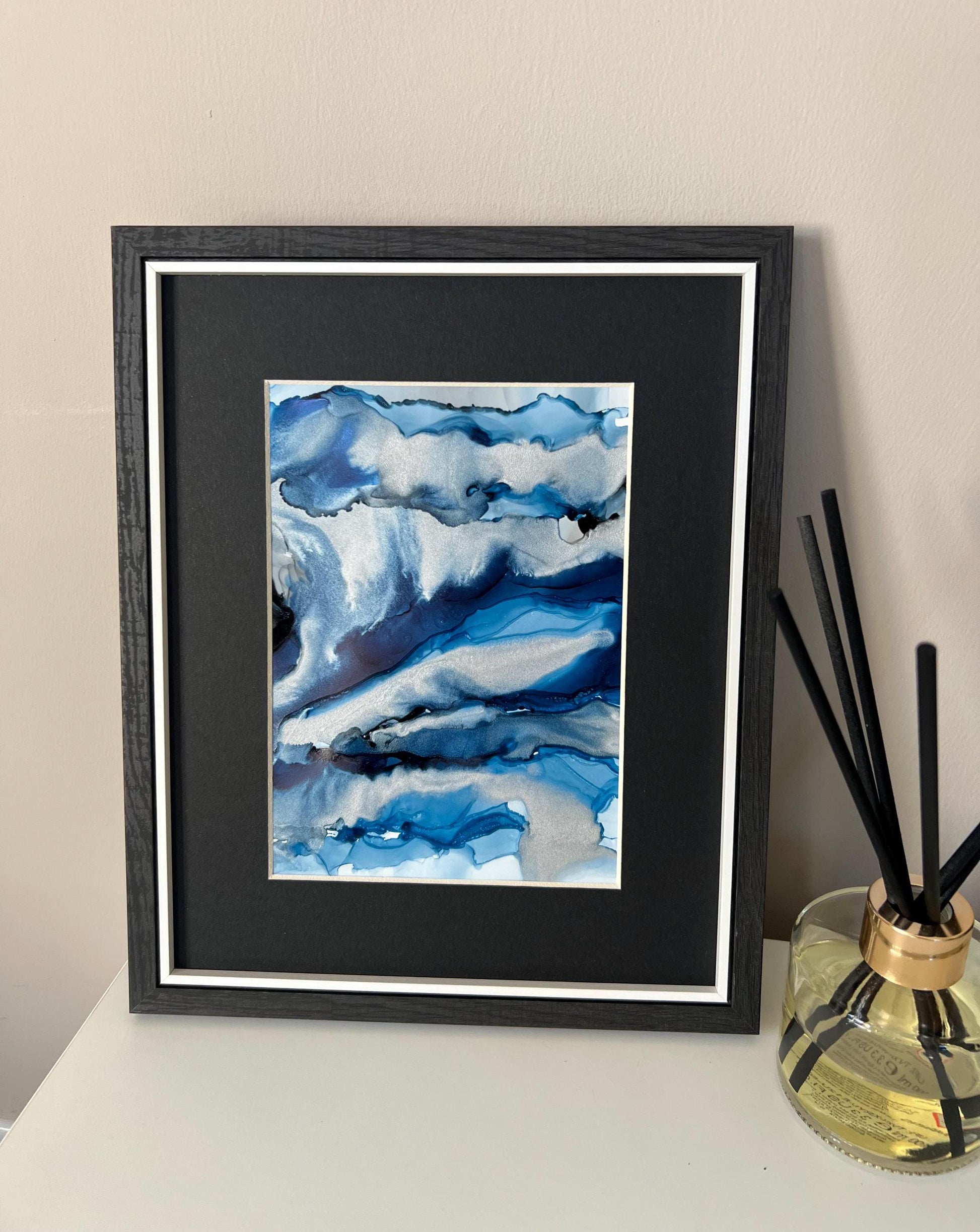 An abstract ink painting in shades of blue and silver, presented in a black frame, displayed on a white surface with a scent diffuser to the side.
