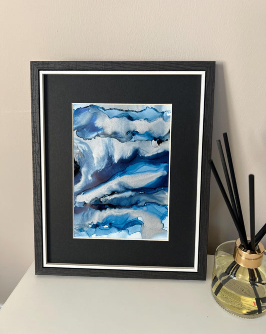 An abstract ink painting in shades of blue and silver, presented in a black frame, displayed on a white surface with a scent diffuser to the side.
