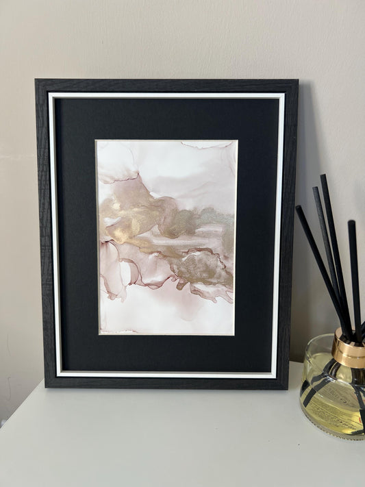 An A5 size contemporary ink painting with pink and gold metallic colors, presented in a black frame, placed on a white surface next to decorative items.