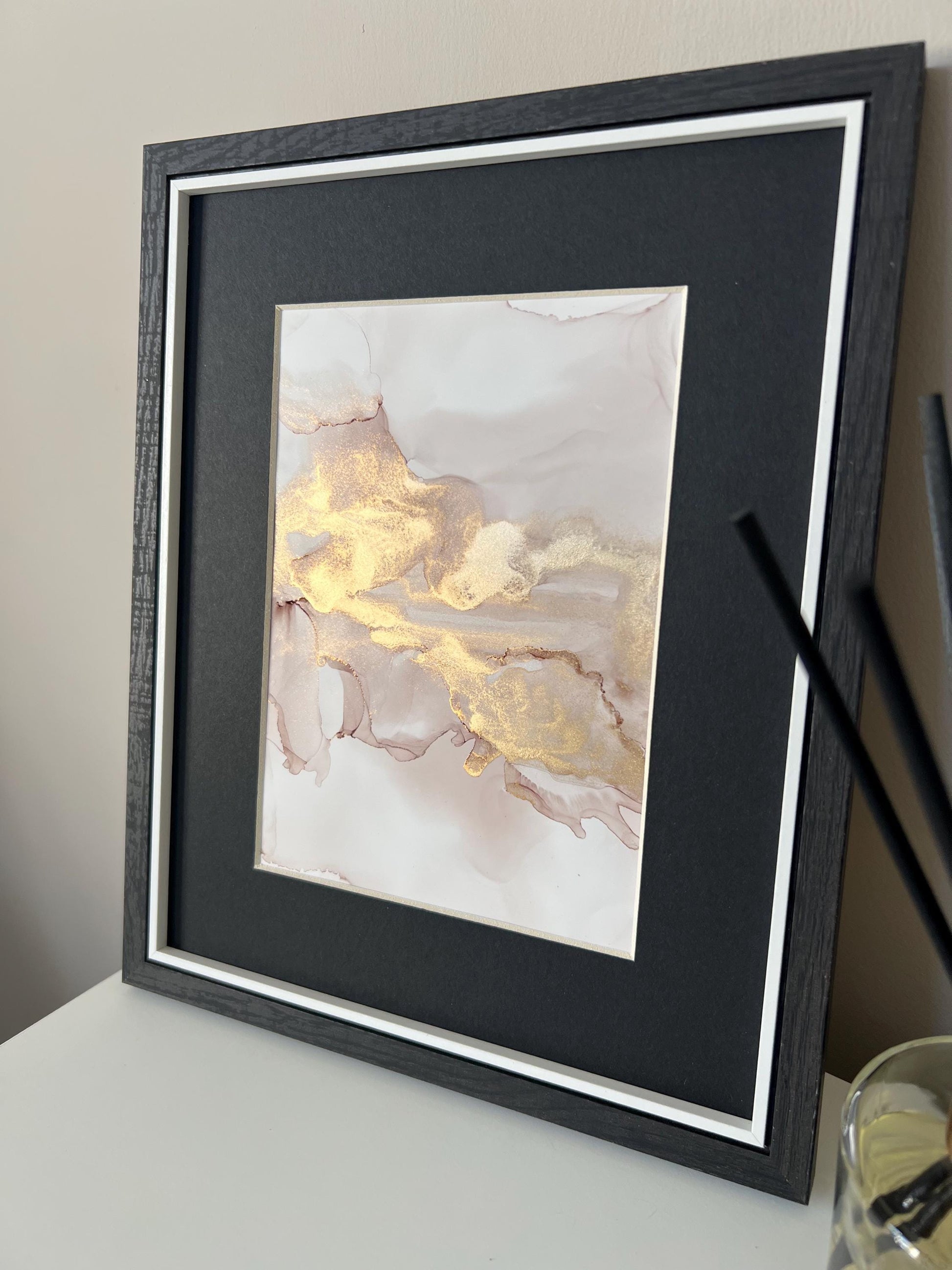 An A5 size contemporary ink painting with pink and gold metallic colors, presented in a black frame, placed on a white surface next to decorative items.