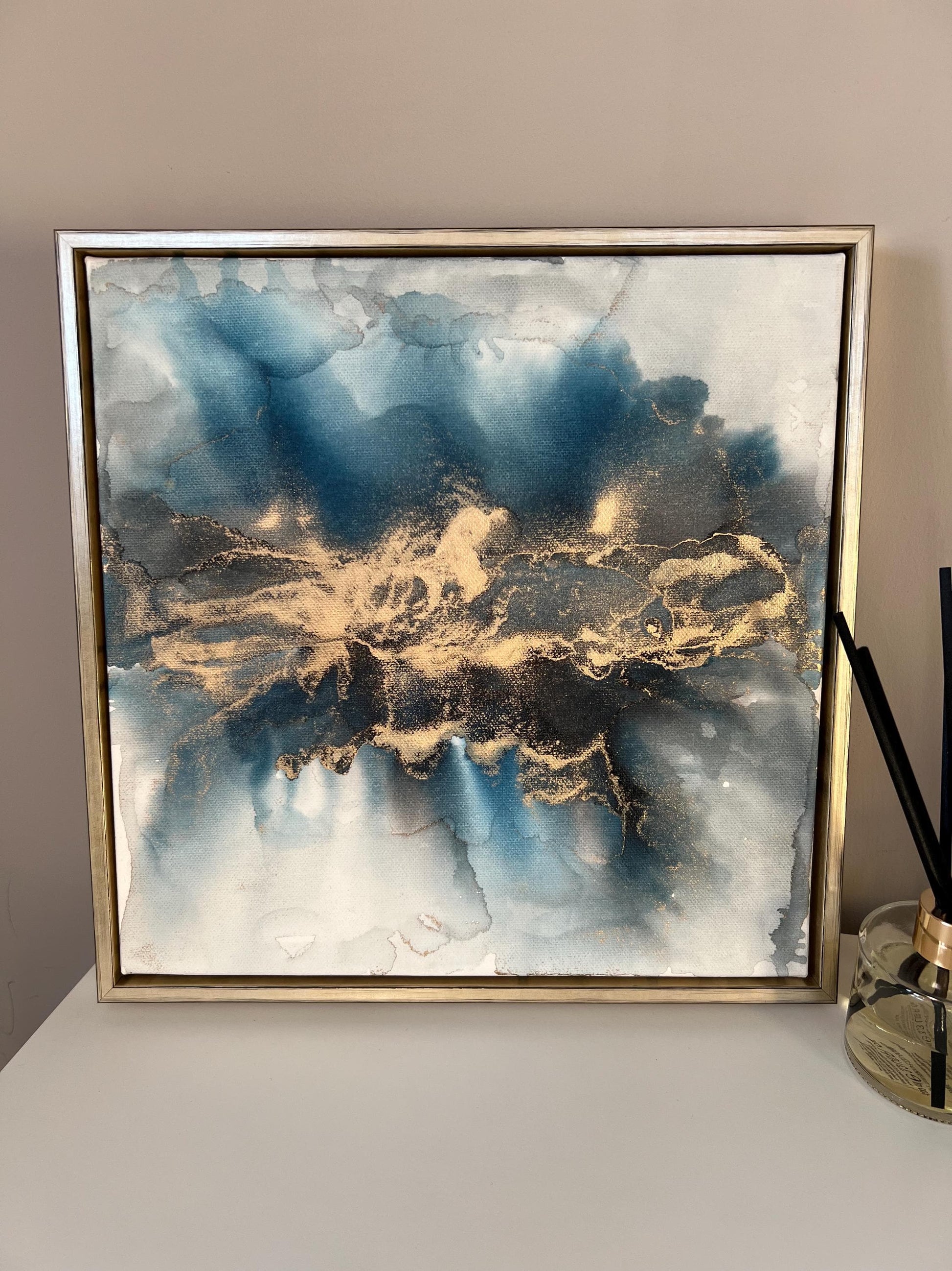 A one-off statement canvas painting featuring an abstract design in alcohol inks with gold and blue colours, with a metallic finish and in a gold finish box frame. 