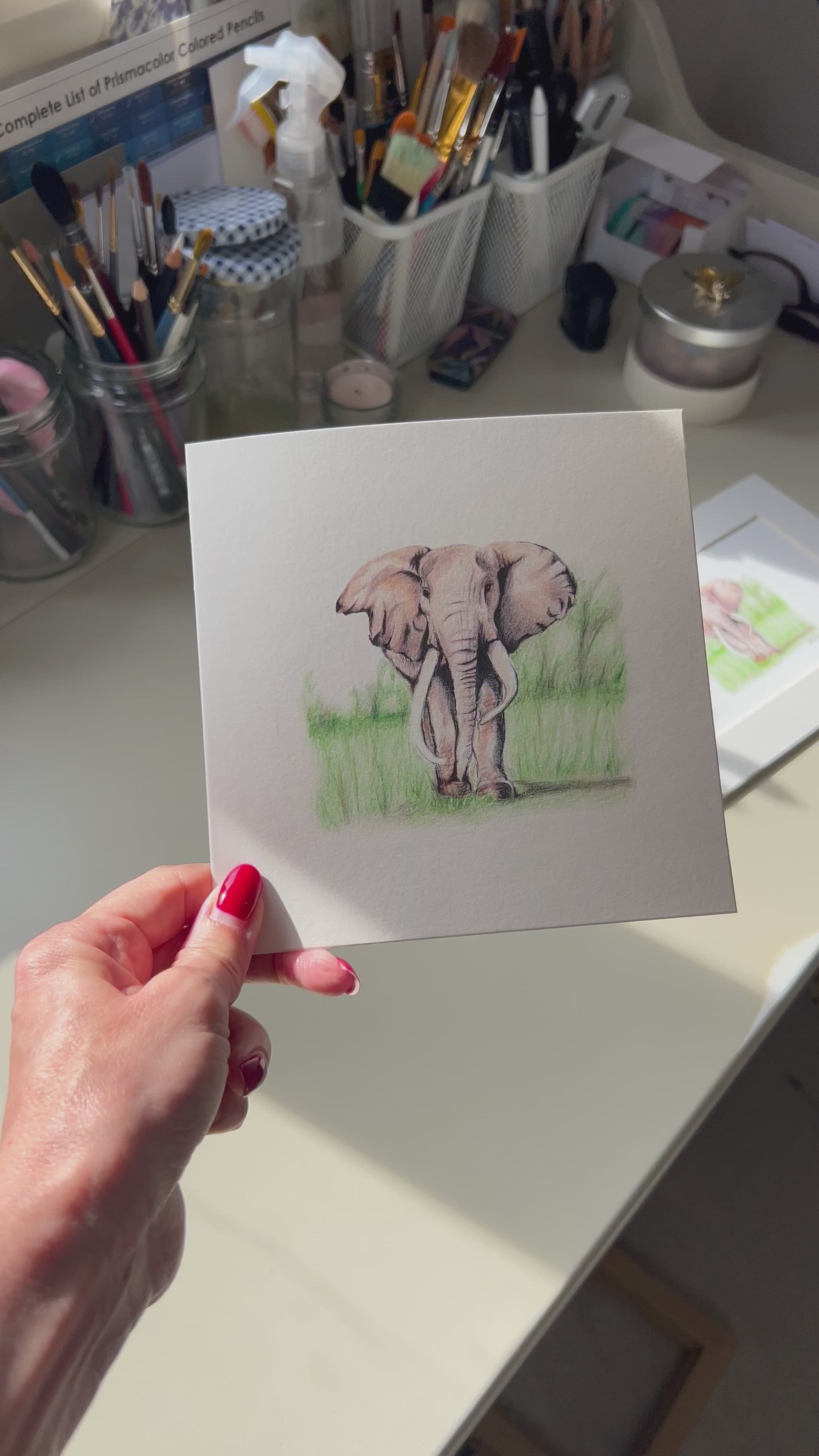 A square greeting card featuring a pencil illustration of an elephant standing in green grass, displayed in a real life art studio. 
