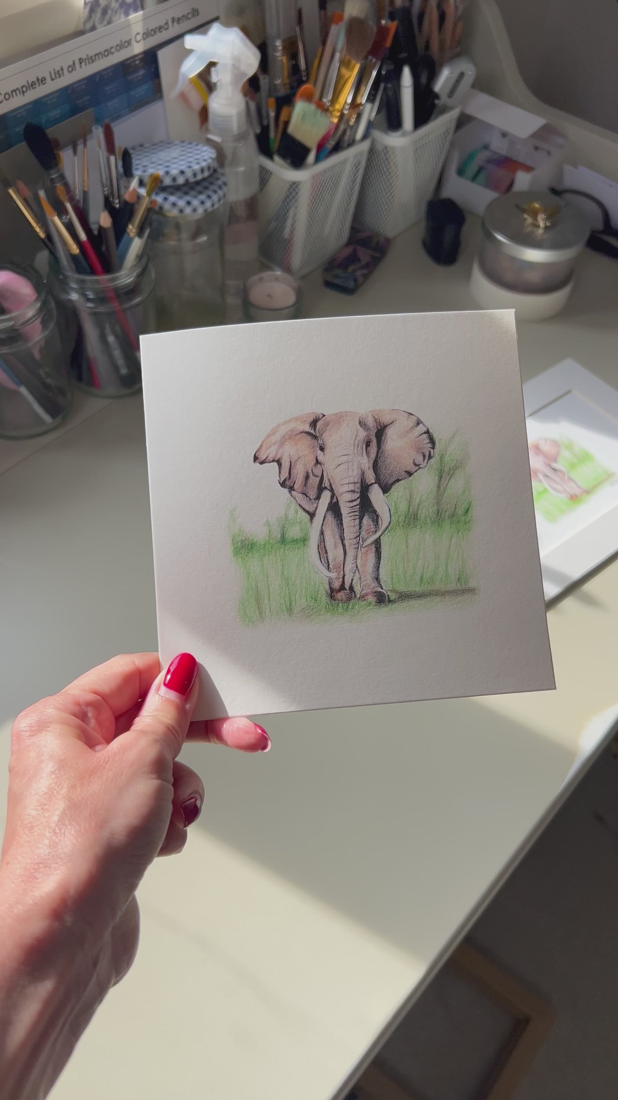 A square greeting card featuring a pencil illustration of an elephant standing in green grass, displayed in a real life art studio. 