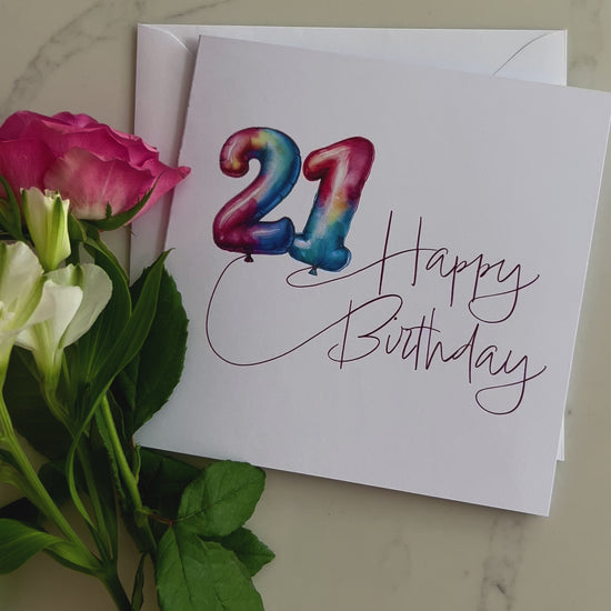 Video of a Birthday card with colourful '21' in balloons and 'Happy Birthday' text, surrounded by flowers on a marble surface.