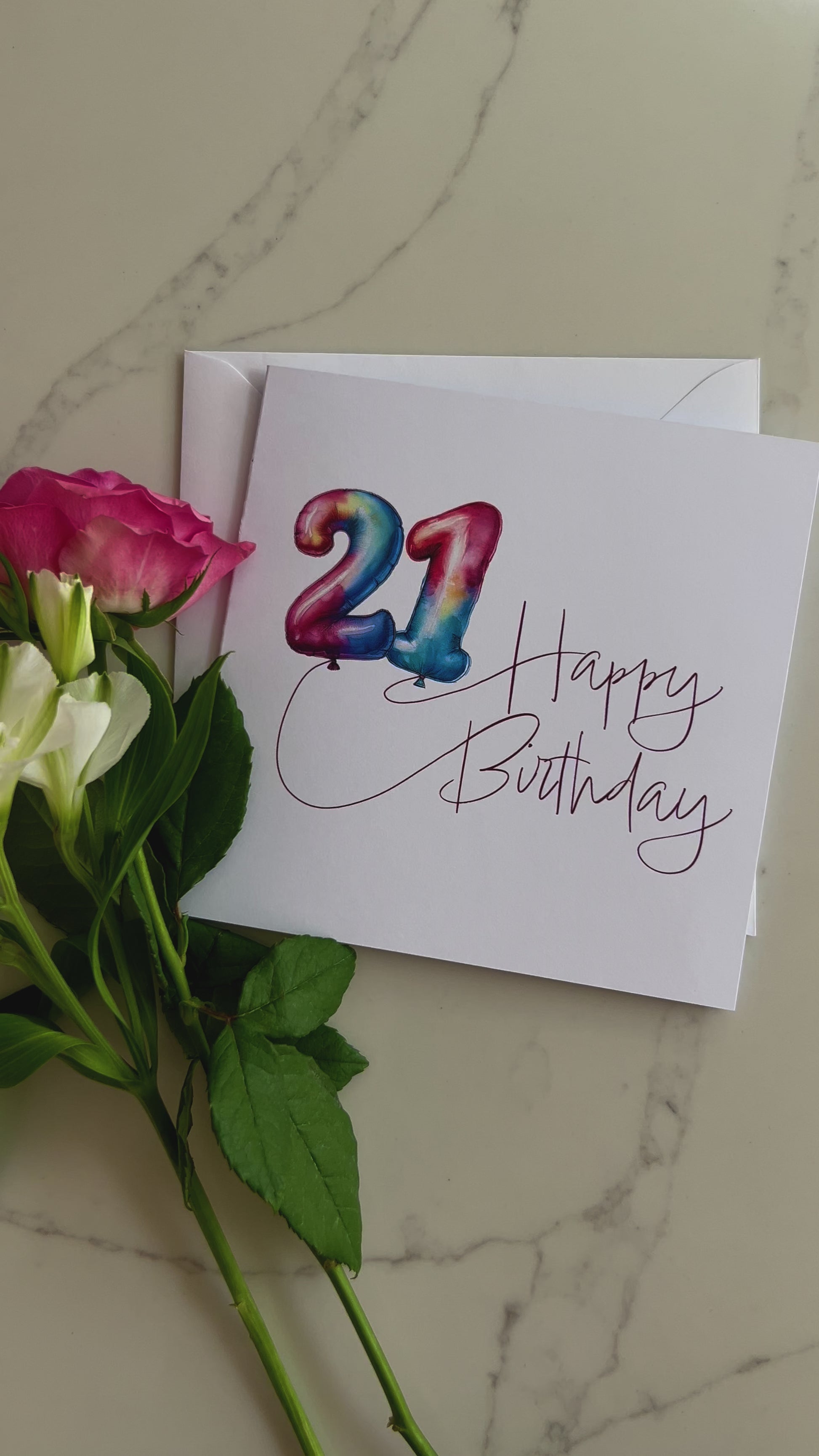 Video of a Birthday card with colourful '21' in balloons and 'Happy Birthday' text, surrounded by flowers on a marble surface.